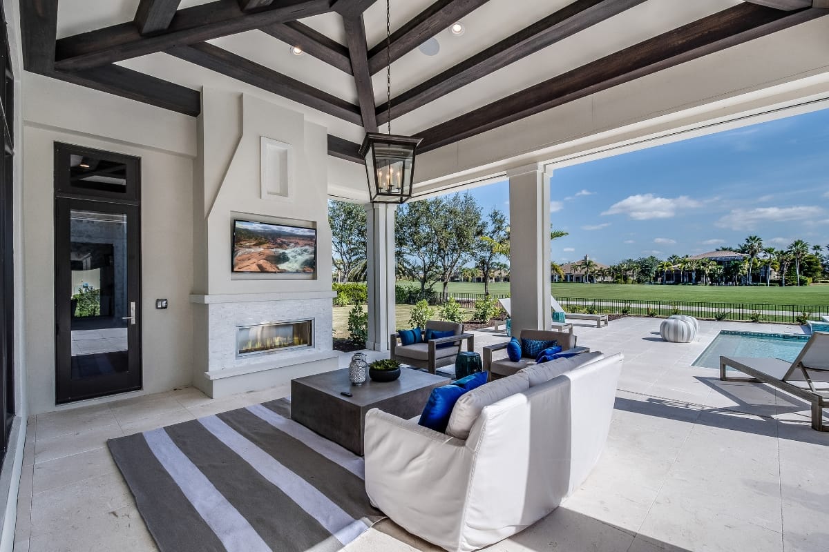 Modern outdoor living area with a white sofa, striped rug, and fireplace, overlooking a pool and golf course. This outdoor smoking lounge features a large open space with wooden ceiling beams and a lantern-style light fixture, creating an inviting atmosphere for relaxation.