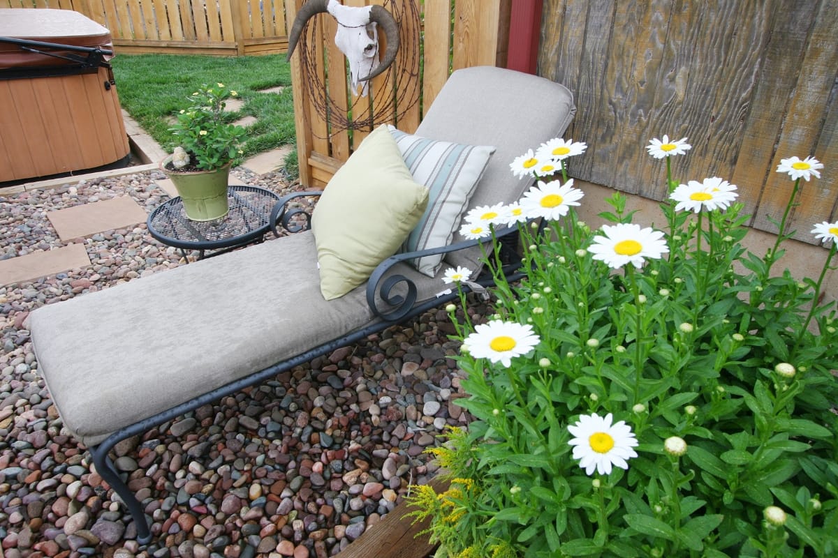 A patio lounge chair with cushions rests on a pebble surface beside a patio table with a potted plant, creating the perfect outdoor smoking lounge. White daisies bloom in the foreground, while a wooden fence and hot tub provide an inviting backdrop.