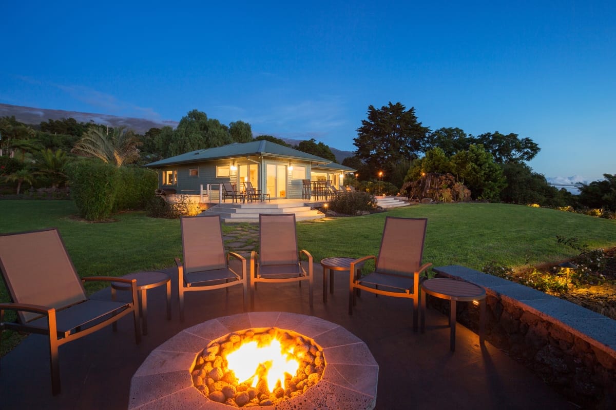 A cozy outdoor fire pit with six chairs creates the perfect setting for an inviting smoking lounge in front of a well-lit house, surrounded by greenery and trees under a clear evening sky.