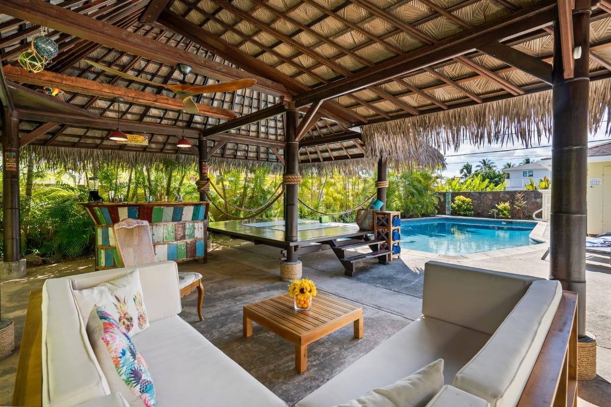 The outdoor patio doubles as an inviting smoking lounge, featuring a thatched roof, wicker furniture, a wooden table adorned with a flower vase, and a cozy hammock. Enjoy views of the sparkling swimming pool framed by swaying palm trees.