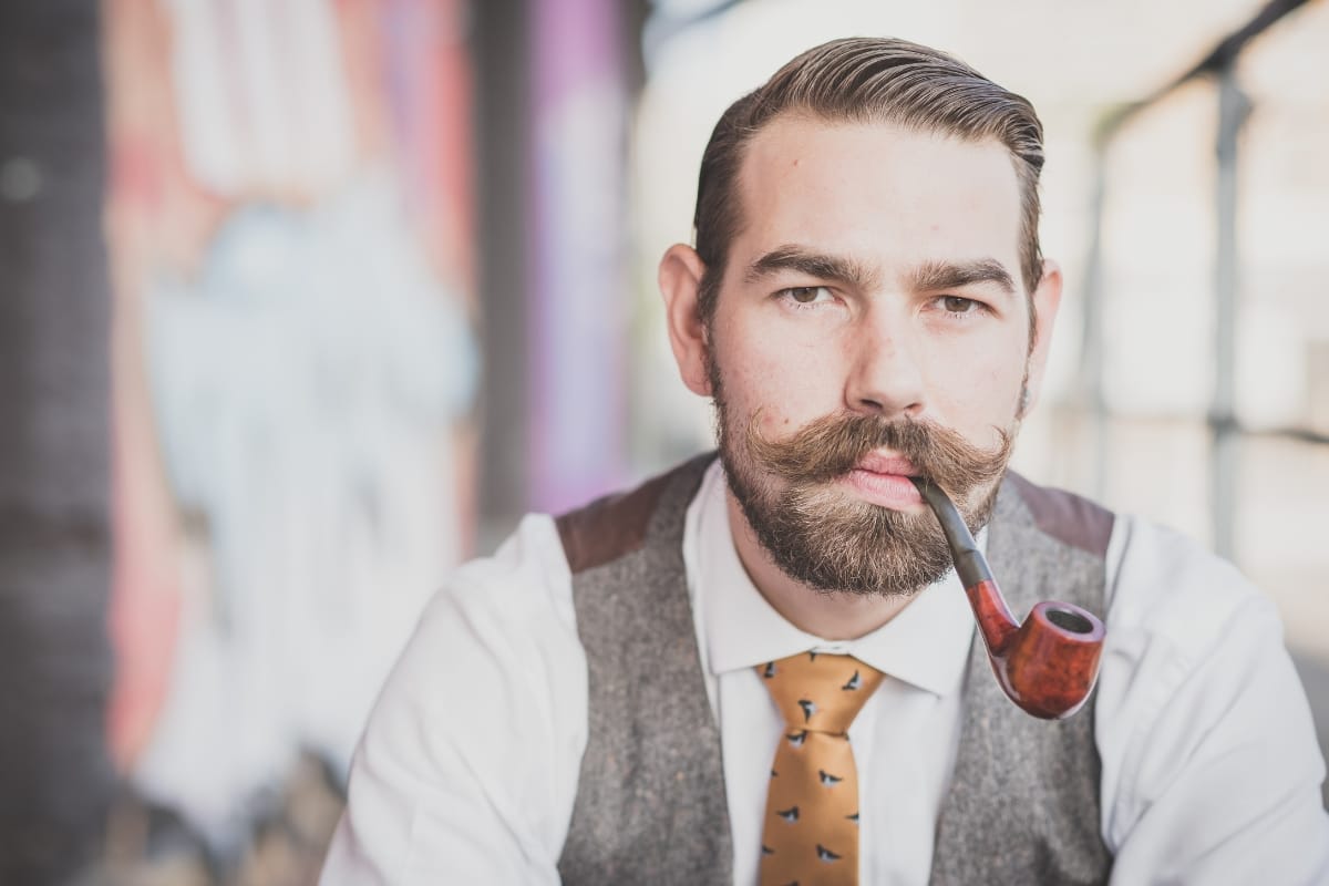 A man with a mustache and beard sits in an outdoor smoking lounge, wearing a vest, tie, and shirt, holding a pipe in his mouth.