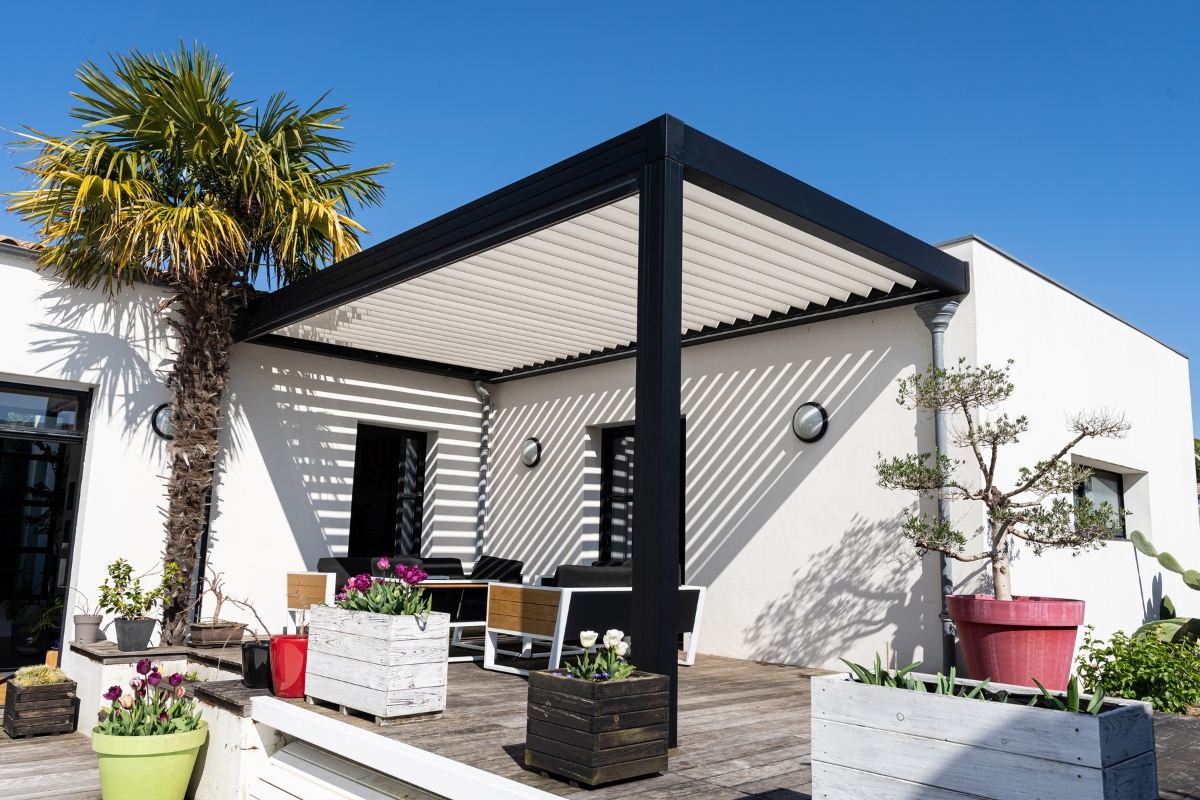 A modern patio serves as an inviting outdoor smoking lounge, featuring a black pergola, wooden furniture, and potted plants, including a palm tree, all set against a clear blue sky.