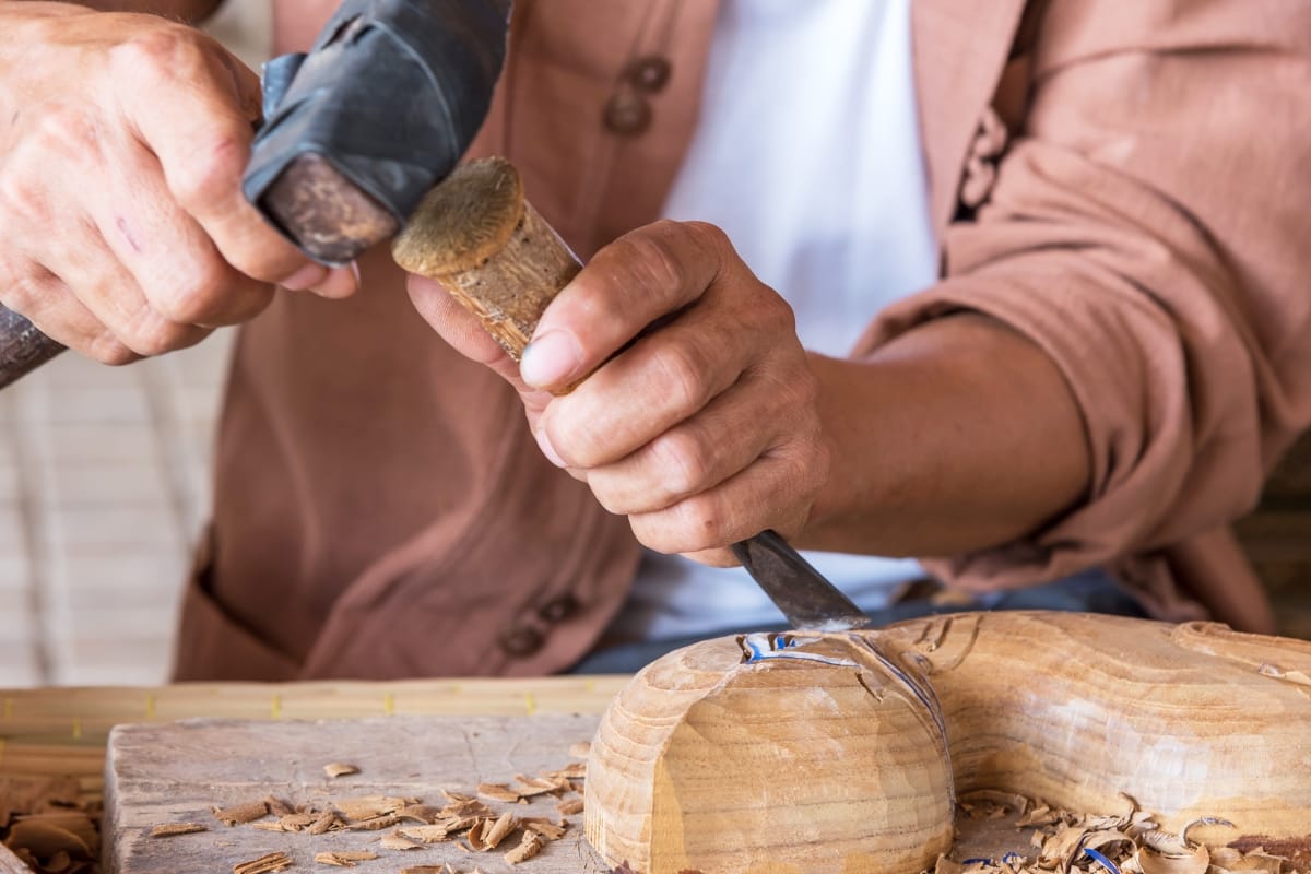 A person meticulously carving wood with a chisel and mallet, focusing on intricate details of a partially shaped piece destined to become one-of-a-kind handcrafted smoking accessories, surrounded by delicate curls of wood shavings.