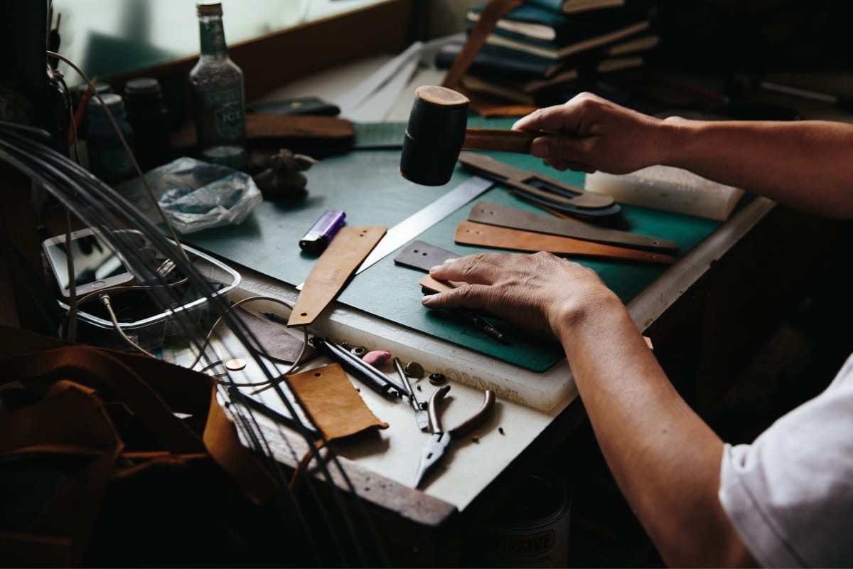 Amidst a cluttered workbench, a person diligently uses a mallet to shape leather pieces, crafting exquisite handcrafted smoking accessories. Tools and materials lie scattered around, telling the story of dedication and artistry.