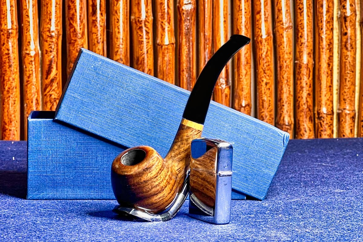 A wooden smoking pipe and a metal lighter are beautifully arranged in front of two blue textured boxes against a bamboo backdrop, showcasing exquisite handcrafted smoking accessories.