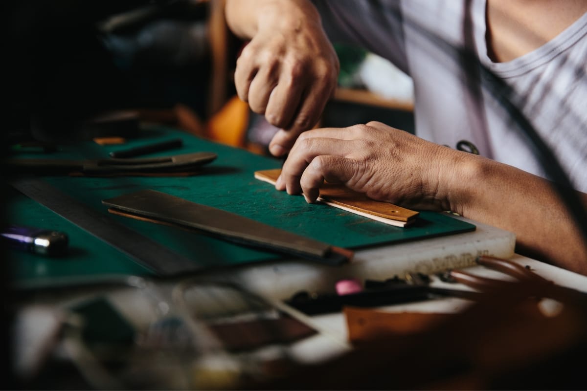 A person meticulously crafting leather on a green workspace, surrounded by various tools and materials, skillfully creates handcrafted smoking accessories.