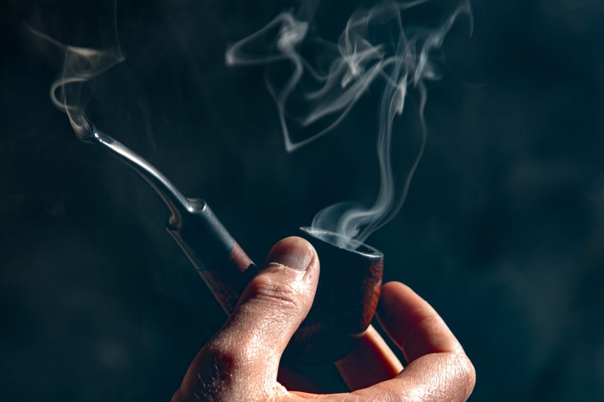 A hand holding a beautifully handcrafted smoking pipe, with visible smoke trails elegantly rising against a dark background.