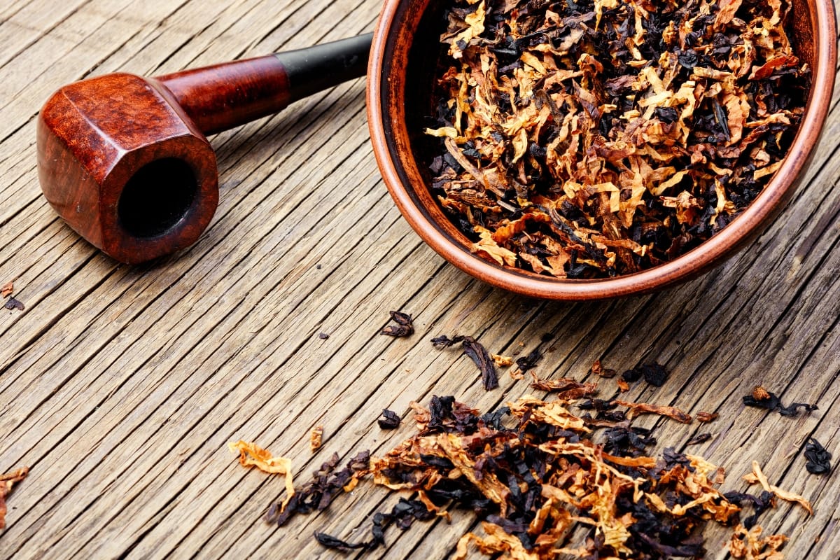 A wooden pipe and a bowl filled with tobacco sit on a wooden surface, accompanied by loose tobacco scattered around, showcasing the art of handcrafted smoking accessories.